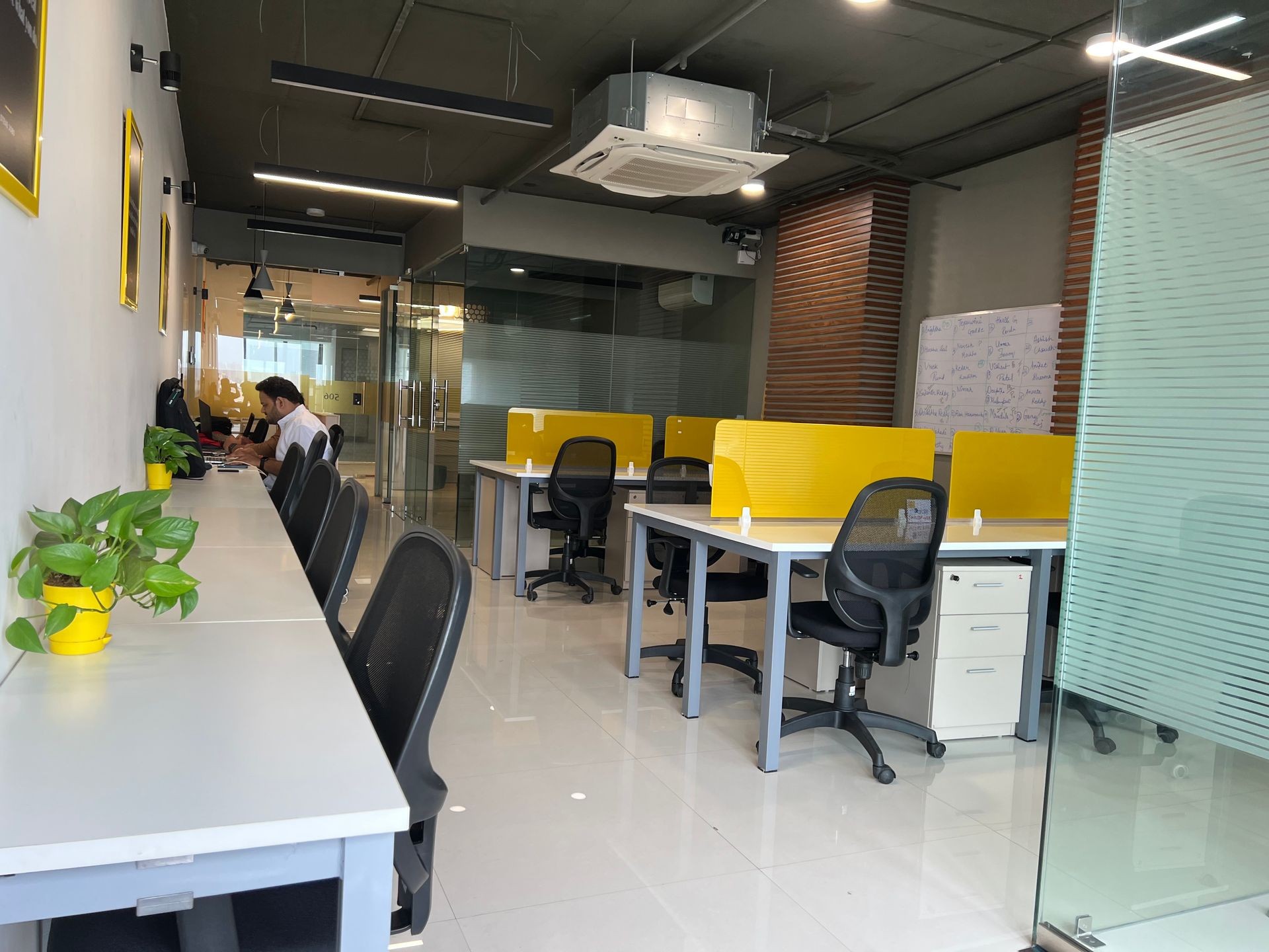 Modern office space with yellow dividers, ergonomic chairs, potted plants, and glass partitions.